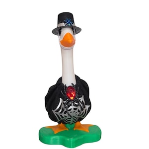 Op de afbeelding: Een wit ganzenbeeldje op een groene voet, met een zwarte hoge hoed met een blauwe spin en een zwarte cape met een zilverkleurig spinnenwebpatroon en een grote rode edelsteen-spin als versiering op de borst.