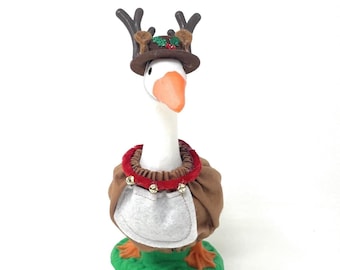 May include: A whimsical goose figurine dressed in a festive reindeer costume. The goose wears a brown hat with antlers, a brown and red collar with bells, and a brown and white apron. It stands on a green base, perfect for holiday decor.