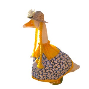 May include: A white goose statue wearing a straw hat with a white flower, long yellow yarn braids, and a yellow dress featuring a blue and white daisy pattern. The dress has a gathered yellow collar and a yellow hem, covering the goose's body.