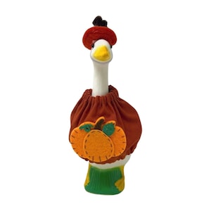 Może przedstawiać: Ceramiczna figurka gęsi w świątecznym stroju. Gęś ma białą szyję i żółty dziób. Jest ubrana w strój w kolorze rdzy z pomarańczową aplikacją z dyni i pasującym kapeluszem, idealnym do jesiennej dekoracji.