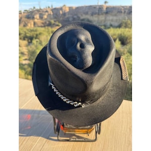May include: Black fedora hat with a skull design on the crown. The hat features a silver chain band and a snakeskin-patterned accent. The hat is displayed on a metal stand.