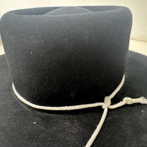 May include: A black hat with a wide brim and a cylindrical crown. A white cord is tied around the base of the crown. The hat is made of a felt-like material.