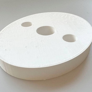 Puede incluir: Base ovalada de plástico blanco con tres agujeros circulares. La base es texturizada y tiene un borde liso y redondeado.
