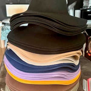 Puede incluir: Una pila de sombreros de ala ancha en varios colores, incluyendo negro, beige, azul marino, lavanda, amarillo y marrón. Los sombreros tienen una forma de fedora clásica, con una copa redondeada y un ala ancha y flexible. Los sombreros están apilados.