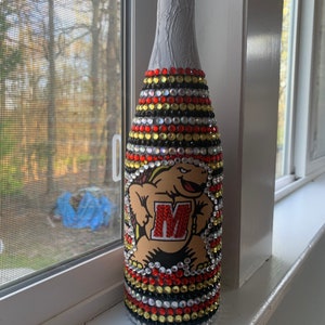 Peut inclure: Une bouteille de champagne blanche décorée de strass noirs, dorés et rouges. La bouteille présente un personnage de dessin animé avec la lettre "M" sur sa poitrine.