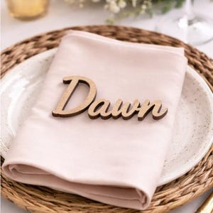 Puede incluir: Una servilleta de tela rosa claro doblada sobre un plato blanco moteado, con una etiqueta de madera cortada a l&aacute;ser que muestra la palabra Dawn en letra cursiva, colocada en el centro de la servilleta como decoraci&oacute;n de mesa.