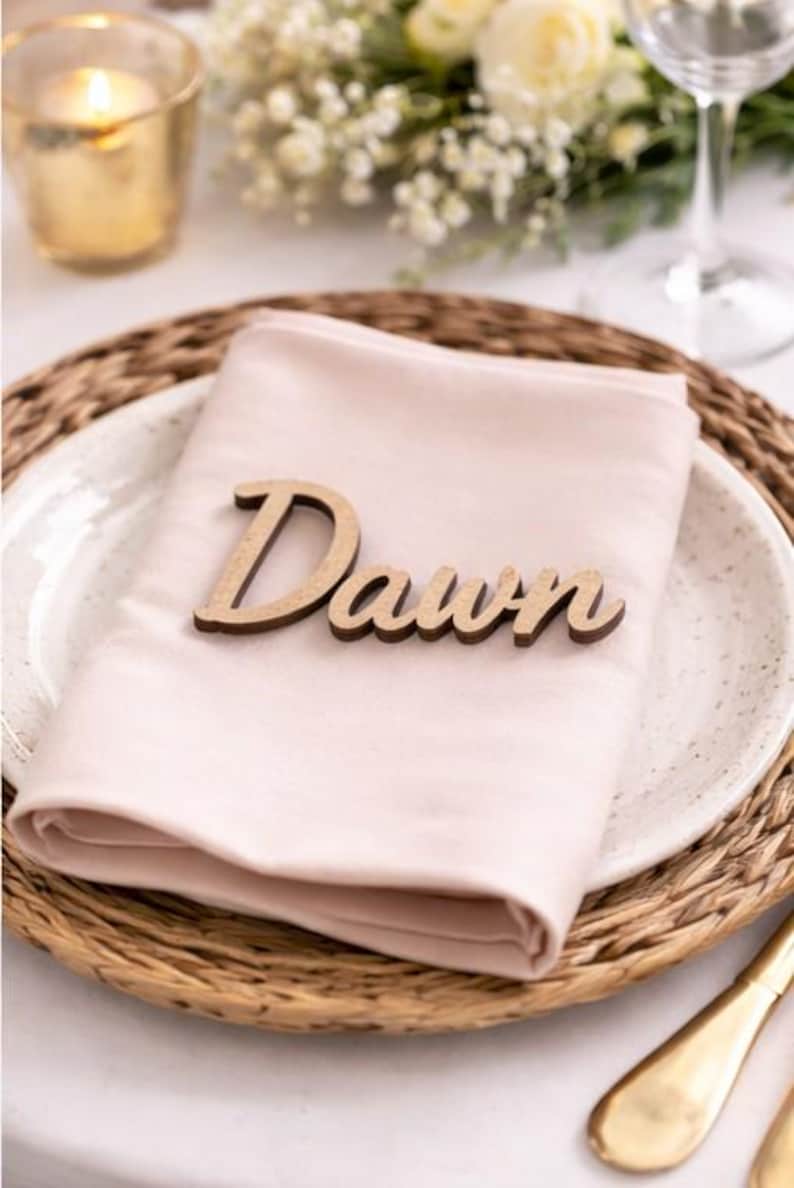 Puede incluir: Una servilleta de tela rosa claro doblada sobre un plato blanco moteado, con una etiqueta de madera cortada a l&aacute;ser que muestra la palabra Dawn en letra cursiva, colocada en el centro de la servilleta como decoraci&oacute;n de mesa.