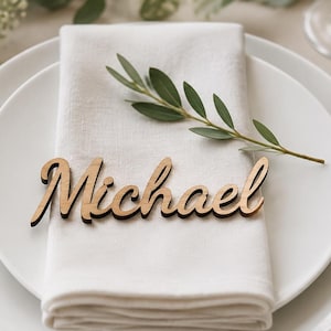 Puede incluir: Un marcador de sitio de madera cortado con l&aacute;ser que presenta el nombre Michael en una fuente cursiva, descansando sobre una servilleta de tela blanca doblada sobre un plato de cer&aacute;mica blanca, acompa&ntilde;ado de cubiertos de metal dorado y una ramita verde.