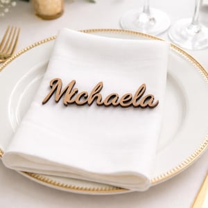 Puede incluir: Una etiqueta de nombre de madera cortada con l&aacute;ser que presenta el nombre Michaela en una fuente cursiva, colocada sobre una servilleta de tela blanca doblada en un plato blanco con borde dorado.