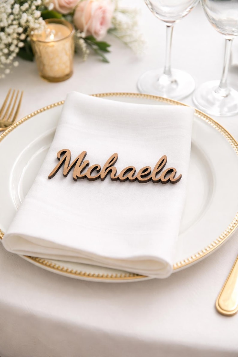 Puede incluir: Una etiqueta de nombre de madera cortada con l&aacute;ser que presenta el nombre Michaela en una fuente cursiva, colocada sobre una servilleta de tela blanca doblada en un plato blanco con borde dorado.