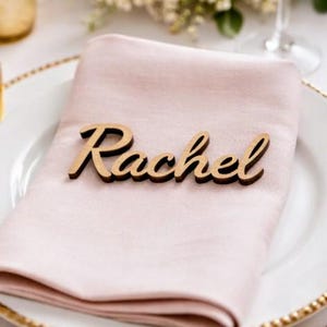 Puede incluir: Una servilleta de tela rosa claro doblada sobre un plato blanco con borde dorado. Sobre la servilleta descansa una etiqueta de madera cortada con l&aacute;ser que muestra el nombre Rachel en letra cursiva con un acabado de veta de madera natural.