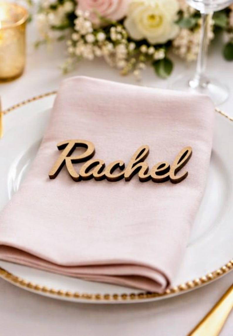 Puede incluir: Una servilleta de tela rosa claro doblada sobre un plato blanco con borde dorado. Sobre la servilleta descansa una etiqueta de madera cortada con l&aacute;ser que muestra el nombre Rachel en letra cursiva con un acabado de veta de madera natural.
