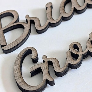 May include: Two laser-cut wooden words in a cursive script font. The top word reads Bride and the bottom word reads Groom. Each word features a light brown wood grain surface with dark, charred edges from the cutting process, creating a layered look.
