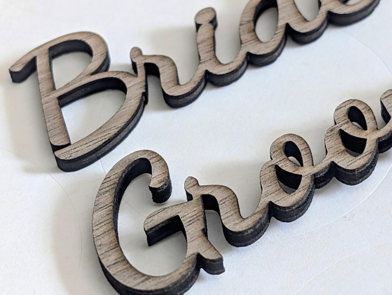 May include: Two laser-cut wooden words in a cursive script font. The top word reads Bride and the bottom word reads Groom. Each word features a light brown wood grain surface with dark, charred edges from the cutting process, creating a layered look.