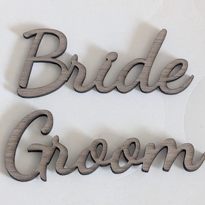 Puede incluir: Dos recortes de madera separados que presentan las palabras Bride y Groom en una fuente cursiva. Las letras tienen un acabado de veta de madera gris oscuro con texturas horizontales visibles en la superficie de cada car&aacute;cter individual.
