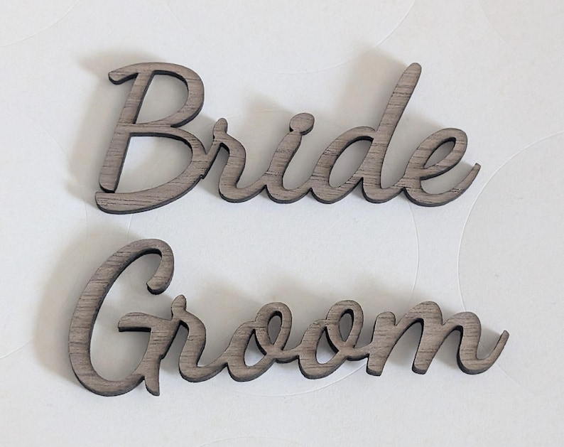 Puede incluir: Dos recortes de madera separados que presentan las palabras Bride y Groom en una fuente cursiva. Las letras tienen un acabado de veta de madera gris oscuro con texturas horizontales visibles en la superficie de cada car&aacute;cter individual.