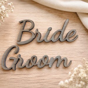 May include: Two dark gray wooden cutouts featuring the words Bride and Groom in a cursive script font. The letters have a smooth finish and are arranged horizontally, with the word Bride positioned above the word Groom on a light wood surface.