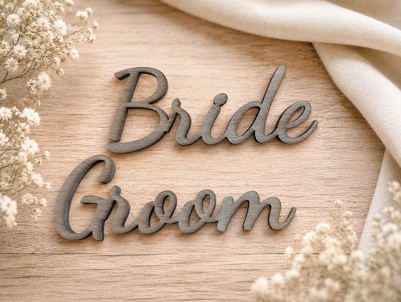 May include: Two dark gray wooden cutouts featuring the words Bride and Groom in a cursive script font. The letters have a smooth finish and are arranged horizontally, with the word Bride positioned above the word Groom on a light wood surface.