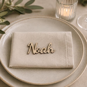 Puede incluir: Una servilleta de tela beige claro doblada sobre un plato de cer&aacute;mica moteada, con una etiqueta de madera cortada a l&aacute;ser que muestra el nombre Noah en letra cursiva. El conjunto incluye un tenedor y un cuchillo dorados, un portavelas y follaje.