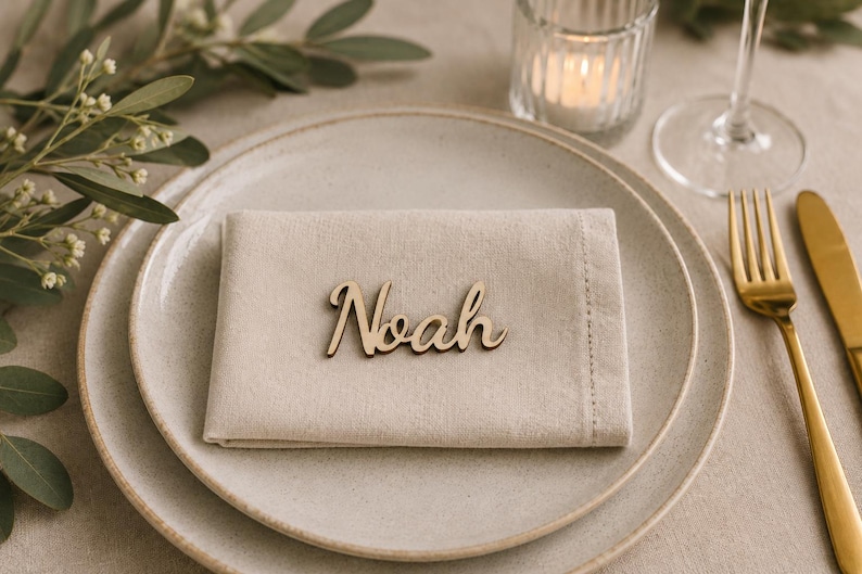 Puede incluir: Una servilleta de tela beige claro doblada sobre un plato de cer&aacute;mica moteada, con una etiqueta de madera cortada a l&aacute;ser que muestra el nombre Noah en letra cursiva. El conjunto incluye un tenedor y un cuchillo dorados, un portavelas y follaje.