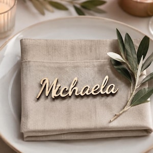 Puede incluir: Una servilleta de lino beige doblada sobre un plato de cer&aacute;mica blanca, con una etiqueta de madera con el texto en cursiva Michaela. Una peque&ntilde;a rama de olivo verde est&aacute; metida en la servilleta, acompa&ntilde;ada de tenedores y un cuchillo dorados.