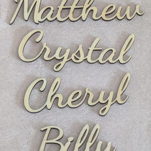 May include: Four individual wooden cutouts featuring the names Matthew, Crystal, Cheryl, and Billy written in a cursive script font. Each name is crafted from light-colored wood with dark edges, displaying a smooth surface and connected lettering.