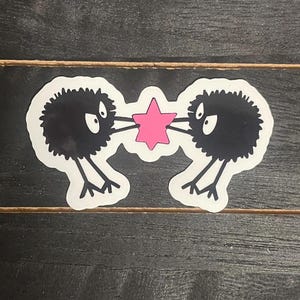 May include: Two black fuzzy creatures with large eyes are holding a pink star. The creatures are facing each other and have a playful expression.