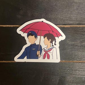 May include: A sticker featuring a cartoon illustration of a boy and girl walking under a red umbrella. The boy is wearing a blue uniform and the girl is wearing a white uniform with a red ribbon.