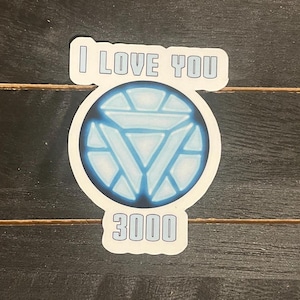 May include: A sticker with the text "I LOVE YOU 3000" in a white, outlined font. The design includes a blue and white circular graphic resembling a reactor core. The sticker is on a dark wood-grain surface.