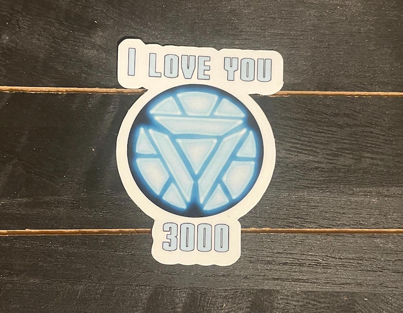 May include: A sticker with the text "I LOVE YOU 3000" in a white, outlined font. The design includes a blue and white circular graphic resembling a reactor core. The sticker is on a dark wood-grain surface.