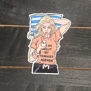 May include: A sticker featuring a cartoon illustration of a woman with blonde hair, wearing an orange t-shirt with the text "Ask Me About My Feminist Agenda." She is holding a drink with a straw. The background has blue and white stripes.