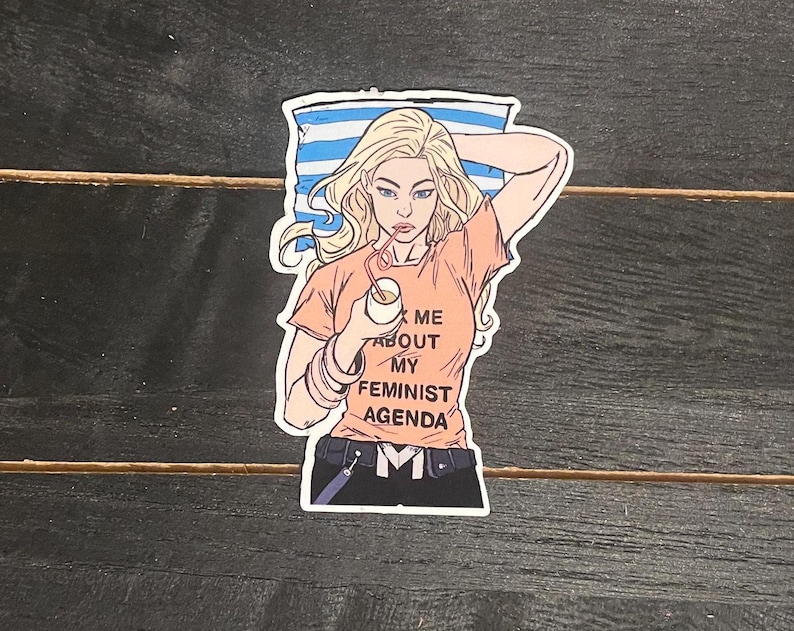 May include: A sticker featuring a cartoon illustration of a woman with blonde hair, wearing an orange t-shirt with the text "Ask Me About My Feminist Agenda." She is holding a drink with a straw. The background has blue and white stripes.