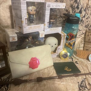 May include: A Harry Potter themed gift set with a variety of items, including a white envelope clutch, a wand, a small wooden box with a golden clock inside, a green wallet with a snake design, a clear water bottle with a Hogwarts crest, a small white figurine of a ghost, a small box with a golden charm, and two small boxes with figurines of Luna Lovegood and Ron Weasley.