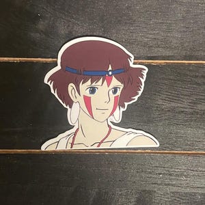 May include: A sticker featuring a cartoon illustration of a young woman with brown hair, wearing a blue headband and a red and white face paint design.