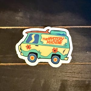 May include: A colorful sticker of the Mystery Machine van from the animated series. The van is light green and yellow with orange and red accents, including flowers and the text "THE MYSTERY MACHINE". A surfboard is on the roof.