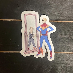 May include: A sticker featuring a cartoon illustration of a superhero in a blue and red suit, standing next to a mirror reflecting a younger version of the same character. The mirror is framed in pink, and a small orange cat is at the superhero's feet.