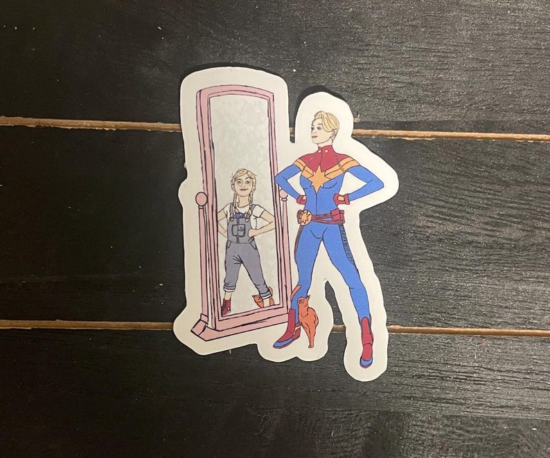 May include: A sticker featuring a cartoon illustration of a superhero in a blue and red suit, standing next to a mirror reflecting a younger version of the same character. The mirror is framed in pink, and a small orange cat is at the superhero's feet.