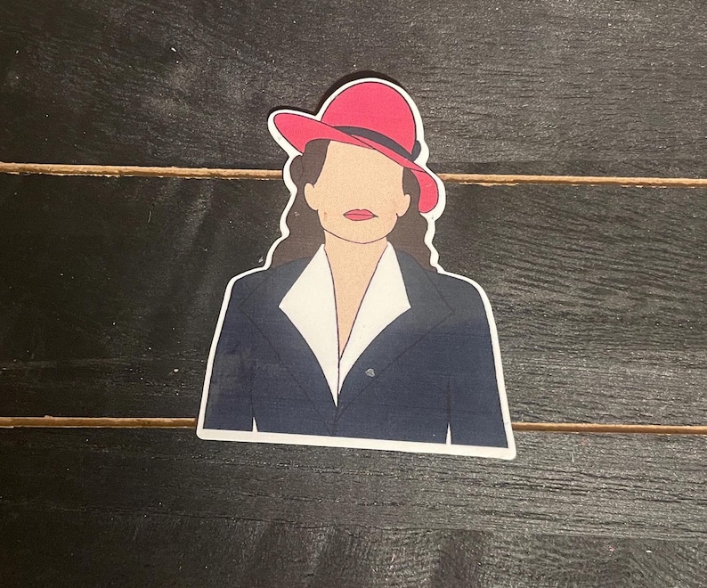 May include: A sticker featuring a stylized illustration of a person wearing a red hat, a navy blue blazer, and a white collared shirt. The sticker is on a dark wood grain surface.