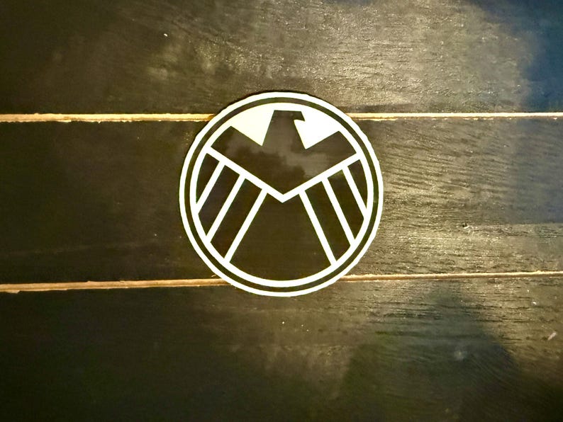 May include: A round, black and white sticker featuring a stylized eagle emblem within a circular border. The design includes a central eagle silhouette and radiating lines, set against a black background.
