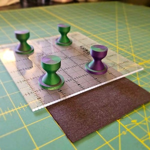 May include: Four green and purple sewing tools on a clear plastic ruler with a grid pattern. The ruler is on top of a brown fabric piece.