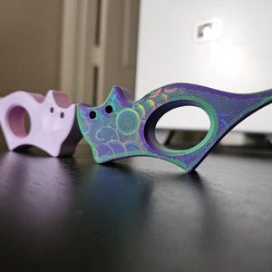 May include: Two cat-shaped rings, one light purple and the other iridescent with green and purple hues. The rings have a circular opening and are placed on a dark surface. The iridescent ring has a pointed tail.