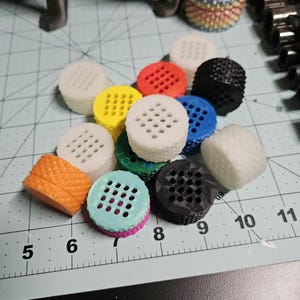 May include: A collection of 11 colorful, round, plastic, 3D printed objects with a grid pattern. The objects are arranged in a semi-circle on a gray surface with white markings.