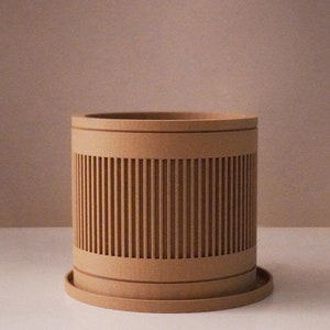 May include: A light brown cylindrical planter with a decorative slatted design. The planter has a matching saucer.