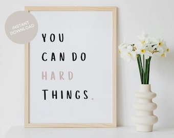 Puede incluir: Una impresión de arte de pared imprimible con el texto "YOU CAN DO HARD THINGS." en blanco y negro con la palabra "HARD" en color rosa claro. La impresión está enmarcada en un marco de madera claro y se muestra sobre una superficie blanca.