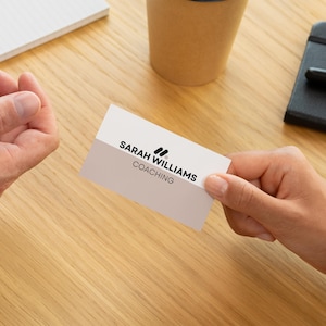 May include: A business card held between two hands, featuring the text "SARAH WILLIAMS COACHING" in black font. The card has a two-tone design with a white top and a light gray bottom. A notepad, a coffee cup, and a small black notebook are also visible.