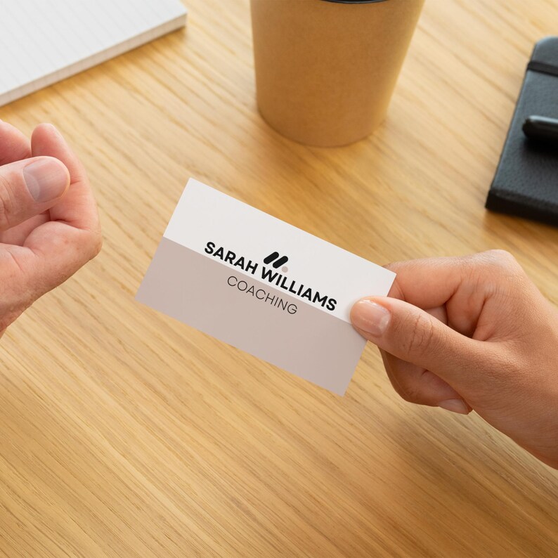 May include: A business card held between two hands, featuring the text "SARAH WILLIAMS COACHING" in black font. The card has a two-tone design with a white top and a light gray bottom. A notepad, a coffee cup, and a small black notebook are also visible.