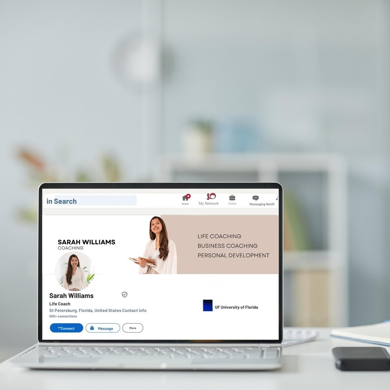 May include: A laptop displays a professional profile page. The profile belongs to Sarah Williams, a life coach. The page highlights "Life Coaching," "Business Coaching," and "Personal Development." The laptop is on a white desk with a notebook and a black device.