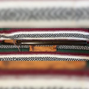 May include: A dark brown wooden flute with a smooth cylindrical body and six finger holes. The instrument features a carved block attached to the top with a leather cord and rests horizontally on a small, rectangular wooden stand.