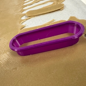 May include: A purple plastic cookie cutter shaped like an oval. The cutter is on top of a sheet of brown cookie dough.