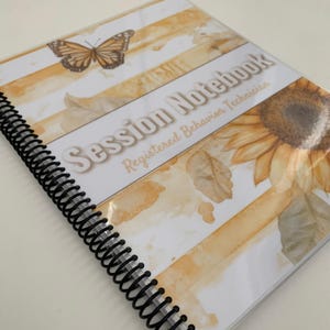 May include: A spiral-bound notebook with a cover featuring a butterfly and sunflower design. The cover has the words "Session Notebook" and "Registered Behavior Technician" printed on it. The background has a watercolor effect with yellow and brown stripes.